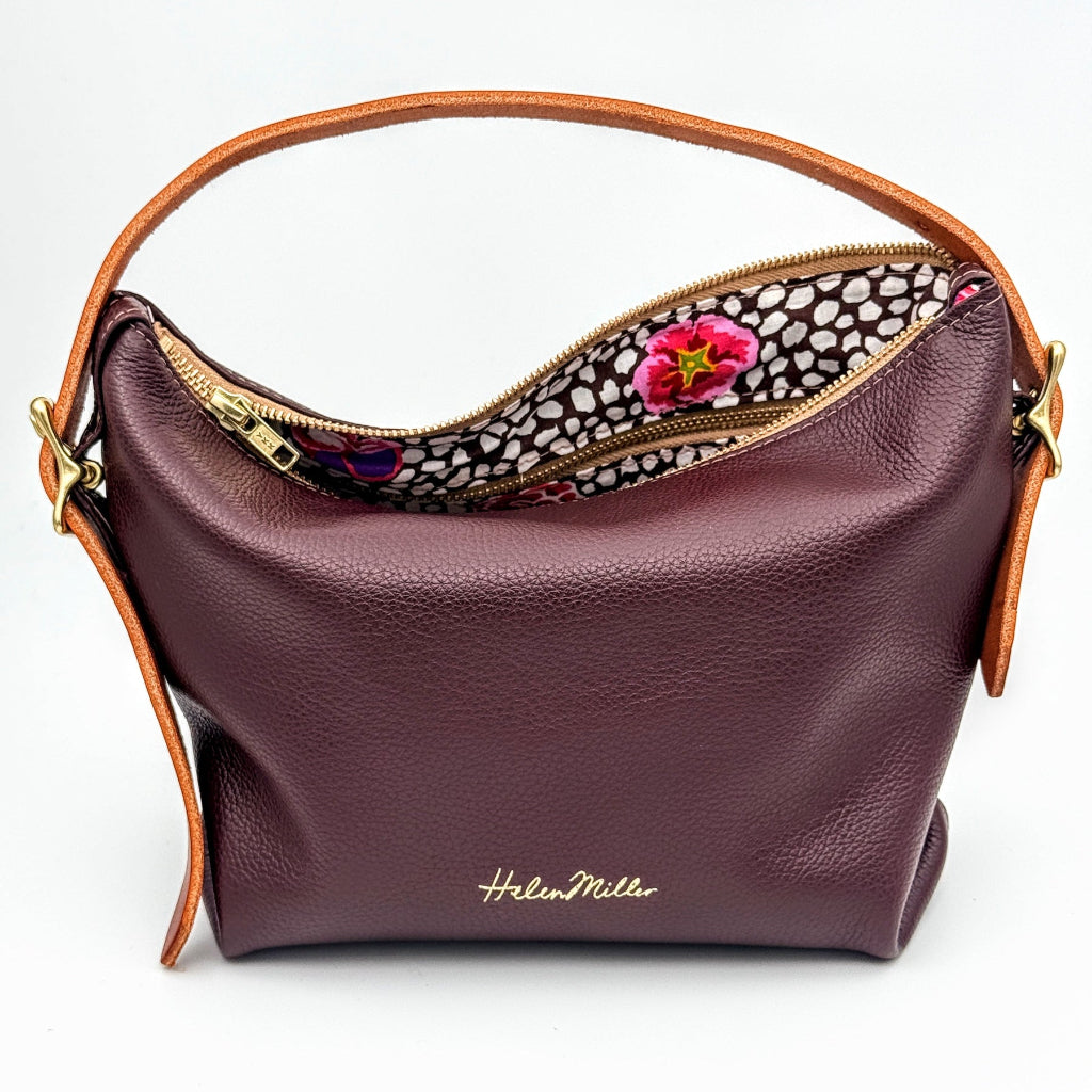 A burgundy pebbled leather hobo shoulder bag with tan leather trim and a gold-tone zipper, shown open to reveal a patterned interior with a pink flower accent. The brand name 'Helmut Miller' is printed in gold on the front.
